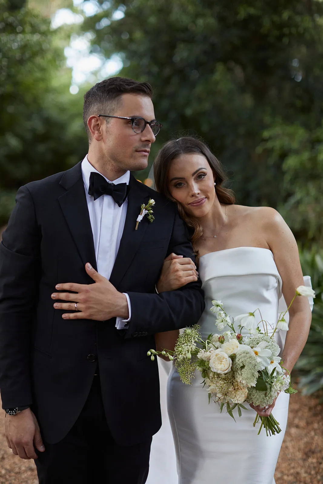 A couple stands together outdoors; the man is dressed in a black tuxedo and glasses, while the woman wears a strapless white bridal gown, holding a bouquet of flowers. He has one arm around her, and they are looking off into the distance with greenery behind them.