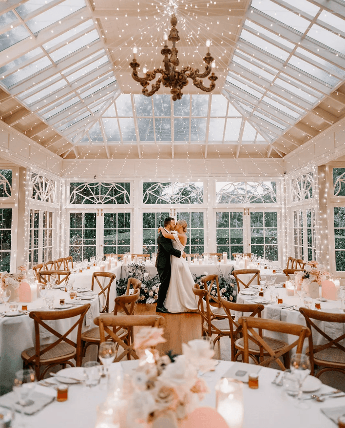 A bride and groom embrace in a romantic dance under cascading string lights in an elegant glass conservatory. The room is filled with neatly arranged tables adorned with soft floral arrangements, candles, and wooden chairs, creating a warm and magical ambiance.