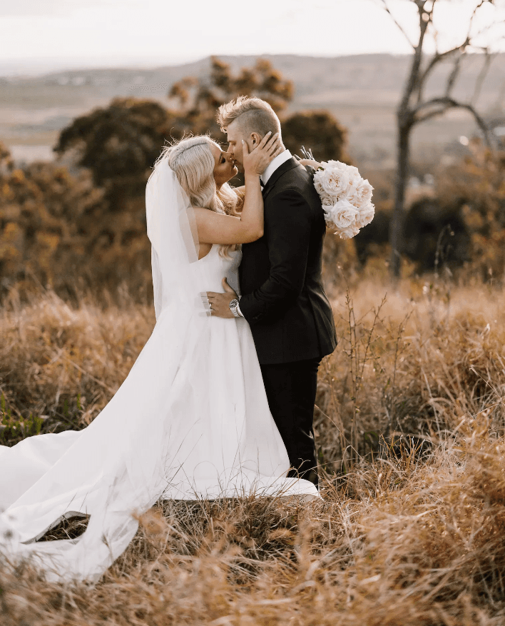 A bride and groom share a kiss in a grassy field with a scenic backdrop of rolling hills and trees. The bride is in a white gown with a long train and veil, holding a bouquet of white flowers. The groom is in a black suit. They are embracing tenderly.