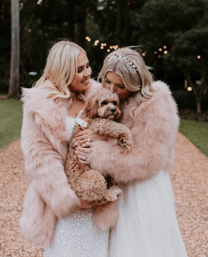 Two brides, both in white dresses and pink faux fur coats, smile and embrace while holding a small, fluffy brown dog. They are outdoors on a wooded path with string lights visible in the background. One bride plants a kiss on the dog's head.
